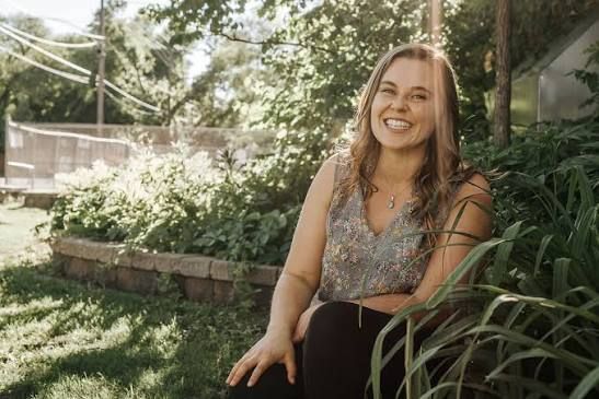 A woman sits in a wooded area, smiling out at the trees.